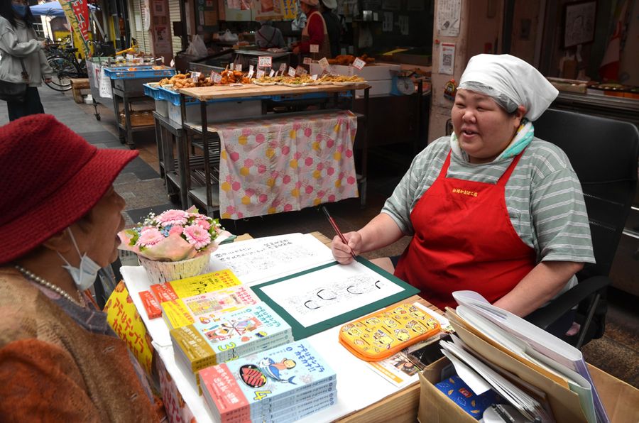 店先で４こまを描く加茂加奈さん。常連客「しげえちゃん」と話しながら。青い本が４巻目（高知市帯屋町２丁目の松岡かまぼこ店）