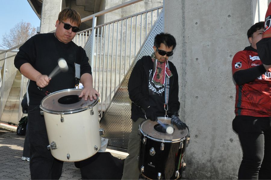 バス待ちでドラムをたたく応援団代表の西森太介さん＝中央＝ら（３月１日）